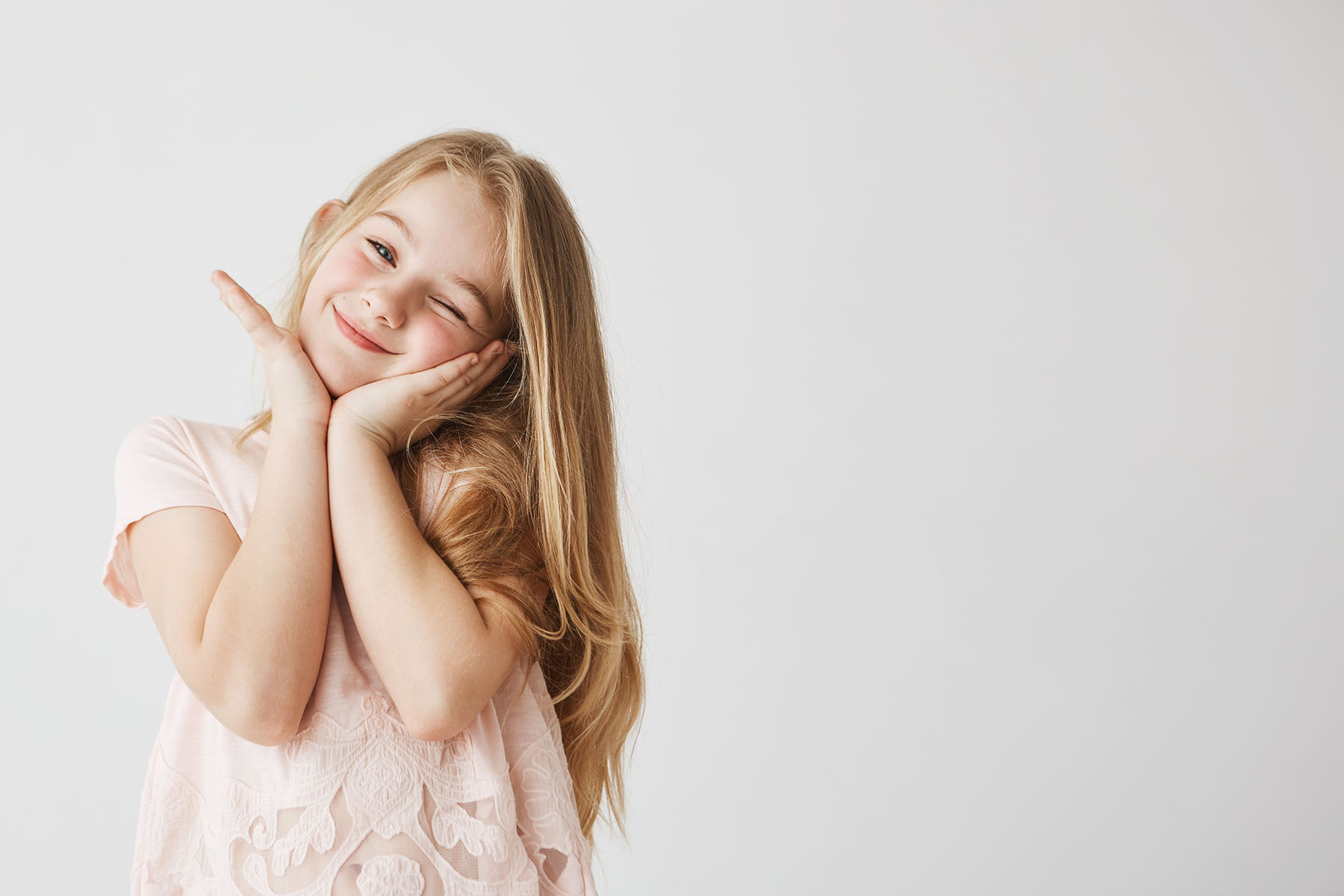 Beutiful little blonde girl smiles at camera winking, posing, touching face with her hands in pink cute dress. Child looking happy and delighted. Copy space.